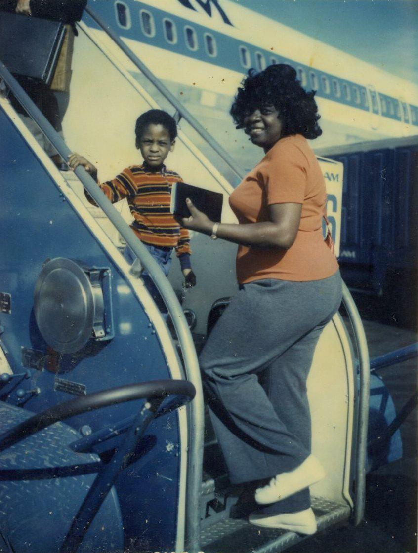 Vie - A vintage photo showing a woman in an orange top and gray pants, smiling while boarding an airplane